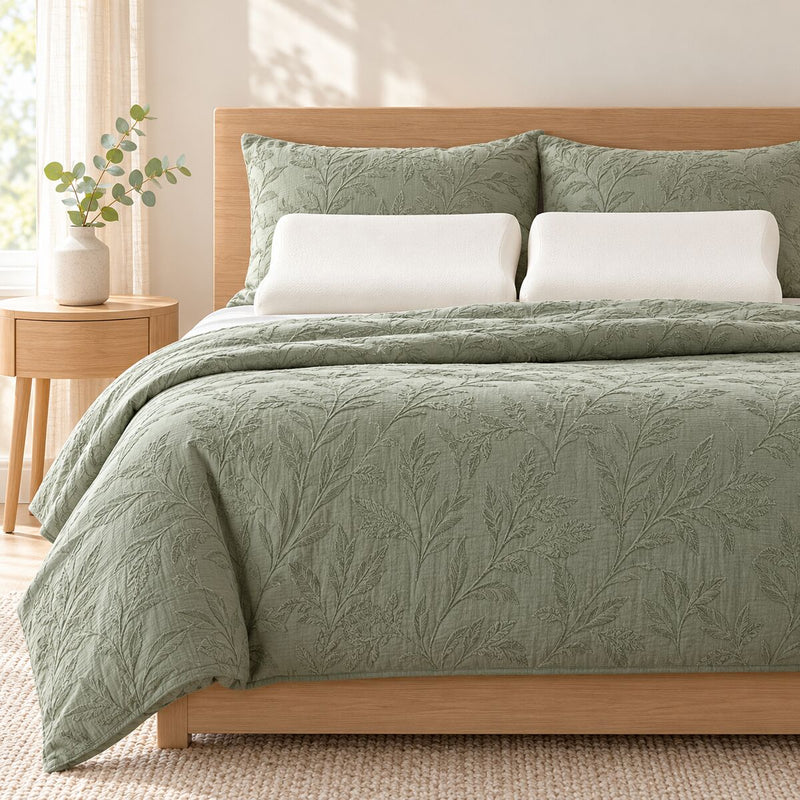 Made bed with a jacquard quilt cover in sage, two memory foam pillows, bedside table with a small artificial eucalyptus stem in a vase.