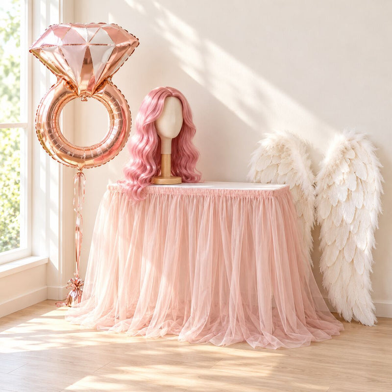 Table with a tulle table skirt, rose-gold diamond-ring foil balloon, a long pink costume wig on a stand and feather fairy wings.